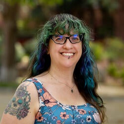 Cierra Covarrubias; Smiling woman with long, wavy green and blue hair, wearing glasses and a floral top. Her tattooed arm is visible. Background is a blurred garden.