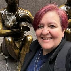 Robin Lee, with short reddish hair smiles for a selfie next to a bronze statue of a seated kangaroo holding a camera.