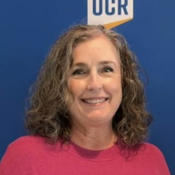 Kelly Jones; Woman with curly hair smiles, standing in front of a blue background with the UCR logo. She wears a pink shirt, conveying a friendly and professional tone.