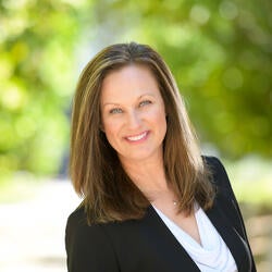 headshot of a JoAnna Van Brocklin  with light brown highlighted hair that comes to her shoulders. She is smiling.he wears a white blouse with a cowl neckline and a small silver necklace. She has on a black blazer. Her arms are down and not visible in the frame, but she seems to be standing in front of greenery that is in a park, blurred, due to a shallow depth of field.